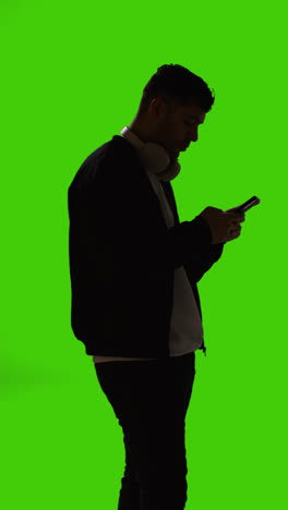 Silueta-De-Vídeo-Vertical-De-Un-Hombre-Con-Auriculares-Inalámbricos-Enviando-Mensajes-De-Texto-En-Un-Teléfono-Móvil-Contra-Una-Pantalla-Verde